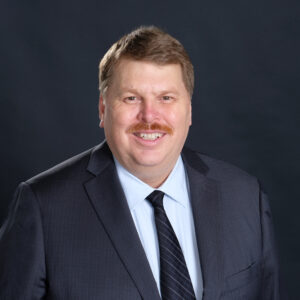 Man in a suit with a moustache, smiling at the camera.