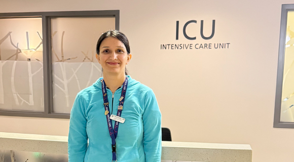 Registered Nurse Mandeep wearing a turquoise long-sleeve shirt, lanyard, and name badge, standing in front of a sign that reads "ICU Intensive Care Unit." She is smiling warmly at the camera.