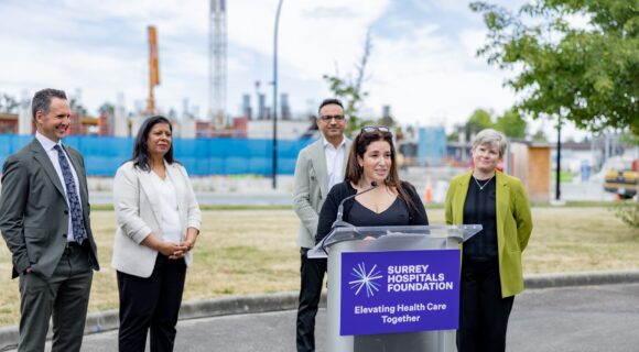 A woman speaks at an outdoor podium with a ‘Surrey Hospitals Foundation: Elevating Health Care Together’ sign, while four people stand behind her smiling. A construction site with cranes and building framework is visible in the background under a partly cloudy sky.
