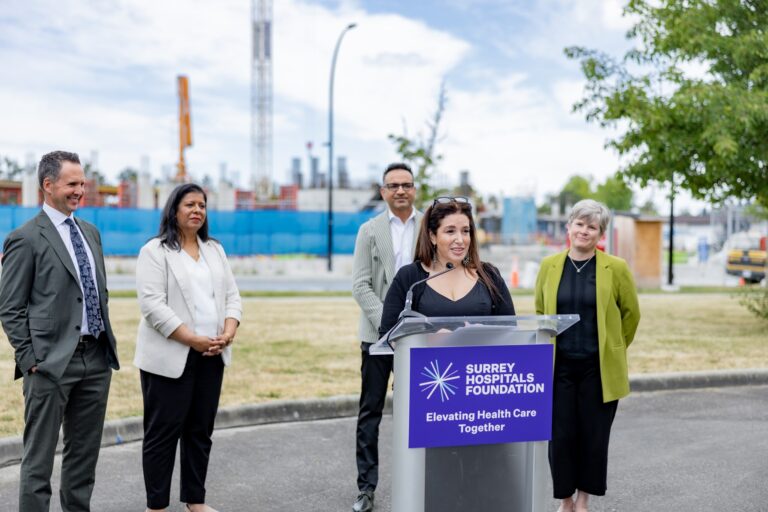 A woman speaks at an outdoor podium with a ‘Surrey Hospitals Foundation: Elevating Health Care Together’ sign, while four people stand behind her smiling. A construction site with cranes and building framework is visible in the background under a partly cloudy sky.