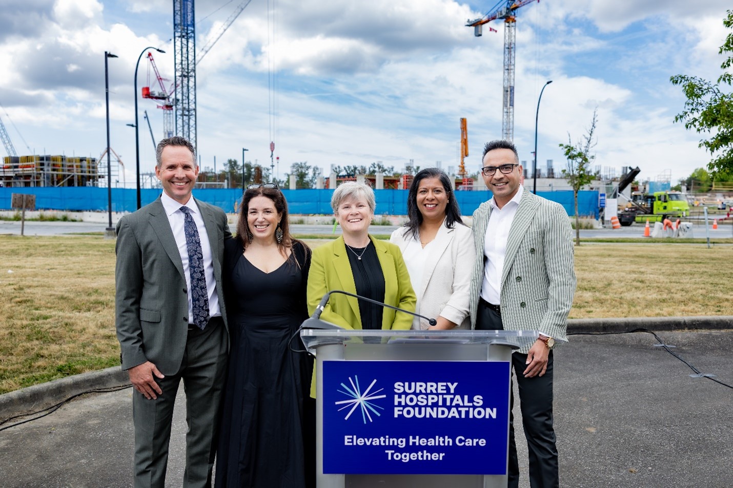 Five people stand smiling behind a podium with a ‘Surrey Hospitals Foundation: Elevating Health Care Together’ sign. A construction site with cranes and building framework is visible behind them under a partly cloudy sky.