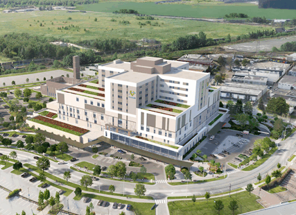 A digital rendering of a large, modern hospital building with multiple stories and green rooftop terraces, surrounded by parking lots, roads, and landscaped areas. The hospital is set against a backdrop of green fields and trees.