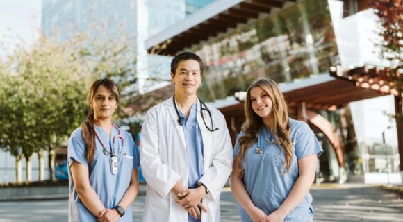 Three smiling healthcare professionals—two women in blue scrubs with stethoscopes and one man in a white lab coat—stand together outside a modern hospital building, with trees and the Surrey Hospitals Foundation logo visible in the background.