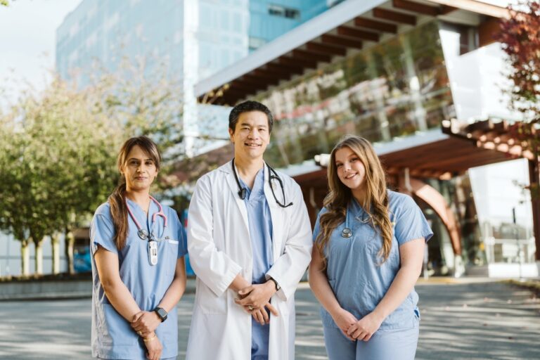 Three smiling healthcare professionals—two women in blue scrubs with stethoscopes and one man in a white lab coat—stand together outside a modern hospital building, with trees and the Surrey Hospitals Foundation logo visible in the background.