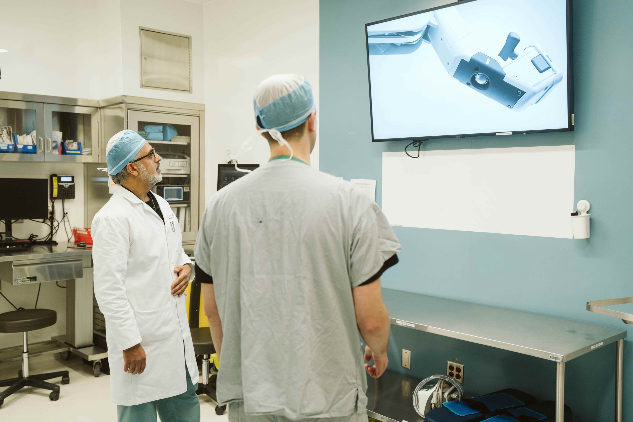 two doctors in a surgery room with one in scrubs and one in a lab coat looking up at a screen with the equipment on it