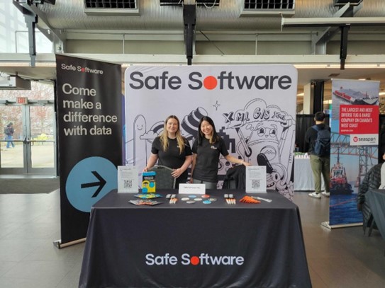 Two women wearing black shirts with a Safe Software logo are smiling behind a table draped in black cloth and a Safe Software logo. They're standing in front of a banner that says "Come make a difference with data"