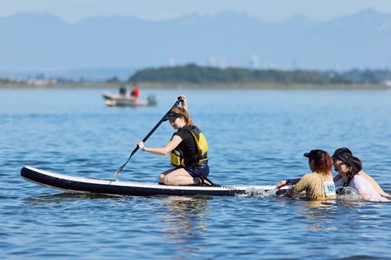 A woman is holding a paddle and she is kneeling on top of a paddle board in the water. Three other persons are in the water pushing the board forward. 