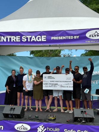 A group of people are standing on top of a stage holding a large cheque. The group is smiling and some are holding their hands up in celebration. 