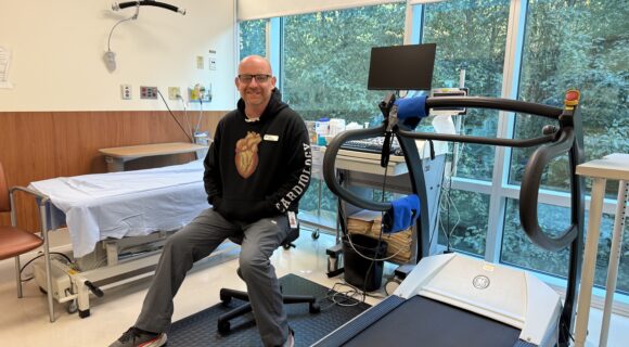 A man is wearing a hoodie with a heart symbol on it. The hoodie has the words "cardiology" written on it. He is sitting down on a stool in a hospital room with a simile on his face.