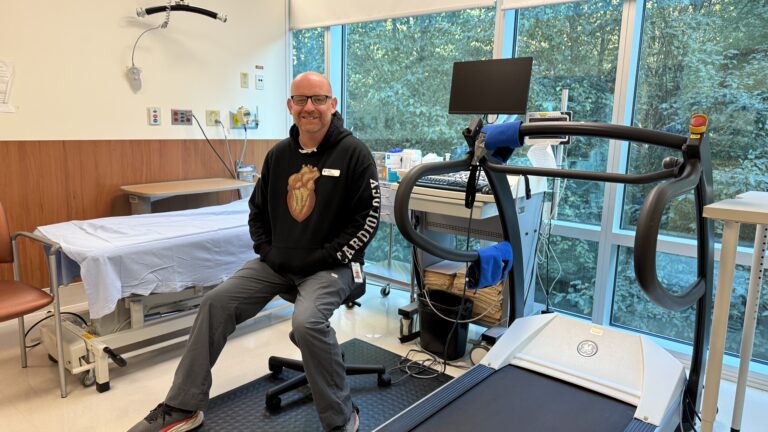 A man is wearing a hoodie with a heart symbol on it. The hoodie has the words "cardiology" written on it. He is sitting down on a stool in a hospital room with a simile on his face.