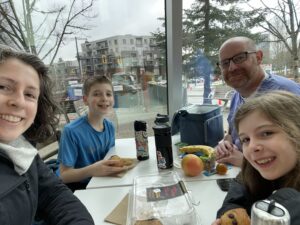 An adults woman, an adult man and two kids are sitting at a lunch table, there is food on the table, they're all looking at the camera smiling.