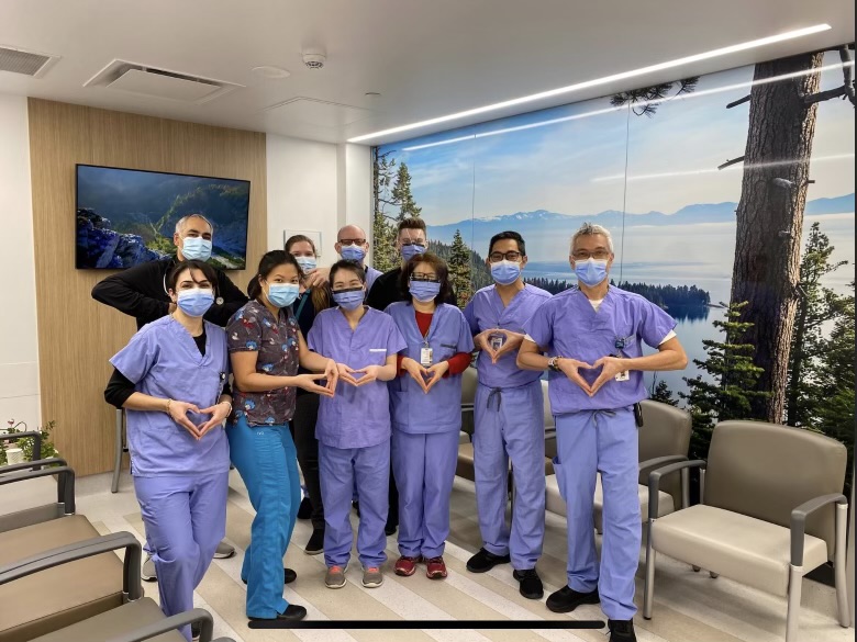 A team of women and men are posing in scrubs with their hands and fingers creating a stylized art - they're wearing masks and smiling warmly with their eyes.