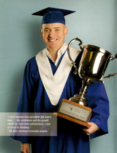A man is in full blue and white graduation gown and cap, holding a large trophy that says "Student of the Year". The man is looking proud, smiling. 