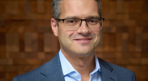 A man with glasses is wearing a full suit posing in front of a wooden wall background. He is looking towards the camera with a soft, professional smile.