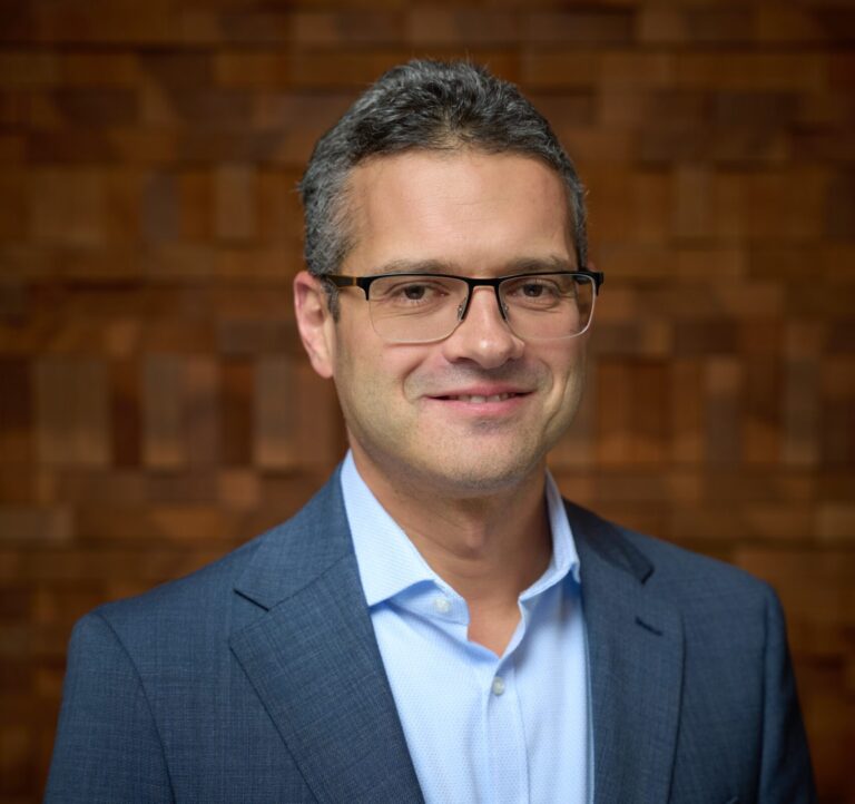 A man with glasses is wearing a full suit posing in front of a wooden wall background. He is looking towards the camera with a soft, professional smile.