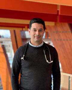 A man wearing a stethoscope and a dark sweater smiling at the camera inside a modern hospital building with wooden beams