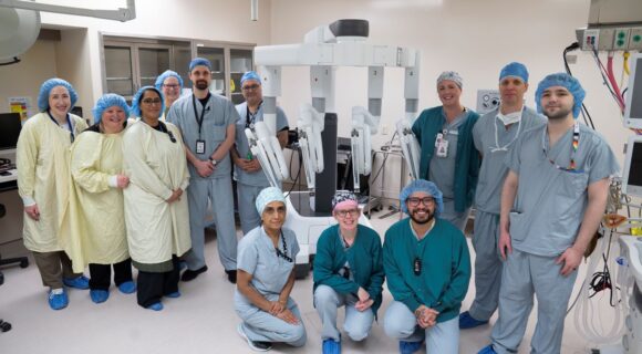 A number of health care staff in scrubs and caps are posing next to the Da Vinci Xi surgical robot. Staff are smiling warmly at the camera.