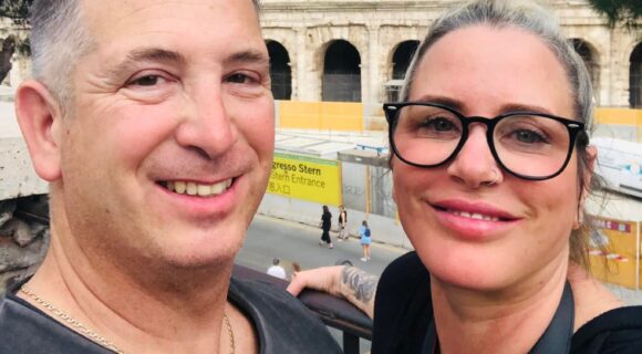 A man and a woman are looking at the camera at a selfie angle, they are both smiling warmly. The colosseum in Rome is in the background.